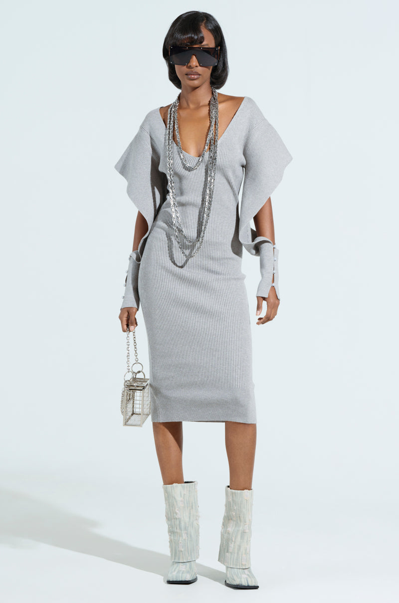 A woman wears the "Off to Brunch" knit midi dress in heather grey, featuring a plunging neckline, paired with silver necklaces and a small silver handbag. She completes her look with wrist accessories, white textured boots, and large black sunglasses.
