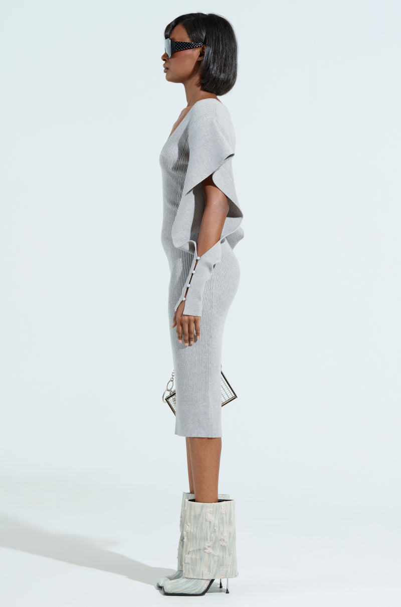 A person poses in profile against a plain backdrop, wearing the OFF TO BRUNCH Knit Midi Dress in heather grey. The dress features asymmetrical sleeves and a flattering fit. They pair it with white layered boots, dark sunglasses, and hold a small geometric handbag.