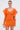 A woman stands against a plain background wearing the OFF TO BRUNCH KNIT MINI DRESS IN ORANGE, featuring dramatic ruffled sleeves and a curve-hugging fit. She accessorizes with gold hoop earrings, a small gold clutch, and has her hair neatly tied back.