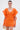 A woman wears the OFF TO BRUNCH KNIT MINI DRESS IN ORANGE with dramatic ruffled sleeves, paired with a gold clutch and hoop earrings. She stands against a plain background with her hair pulled back.