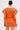 A woman with her hair in a neat bun faces away, wearing the OFF TO BRUNCH KNIT MINI DRESS IN ORANGE—an orange, curve-hugging mini dress with sculpted sleeves. She holds a clear handbag against a plain white background.