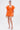 A woman wears the OFF TO BRUNCH KNIT MINI DRESS IN ORANGE, featuring a plunging neckline and open sleeves, styled with gold hoop earrings, gold ankle-strap heels, and a clear clutch against a white background.