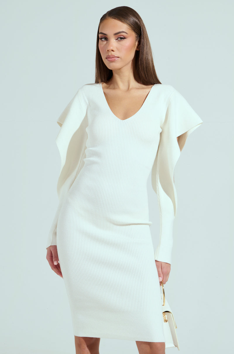 A person in the OFF TO BRUNCH KNIT MIDI DRESS, featuring a fitted white midi design with long sleeves and a V-neckline, stands against a plain background. They're holding a small white handbag and have straight, long hair for an elegant touch.