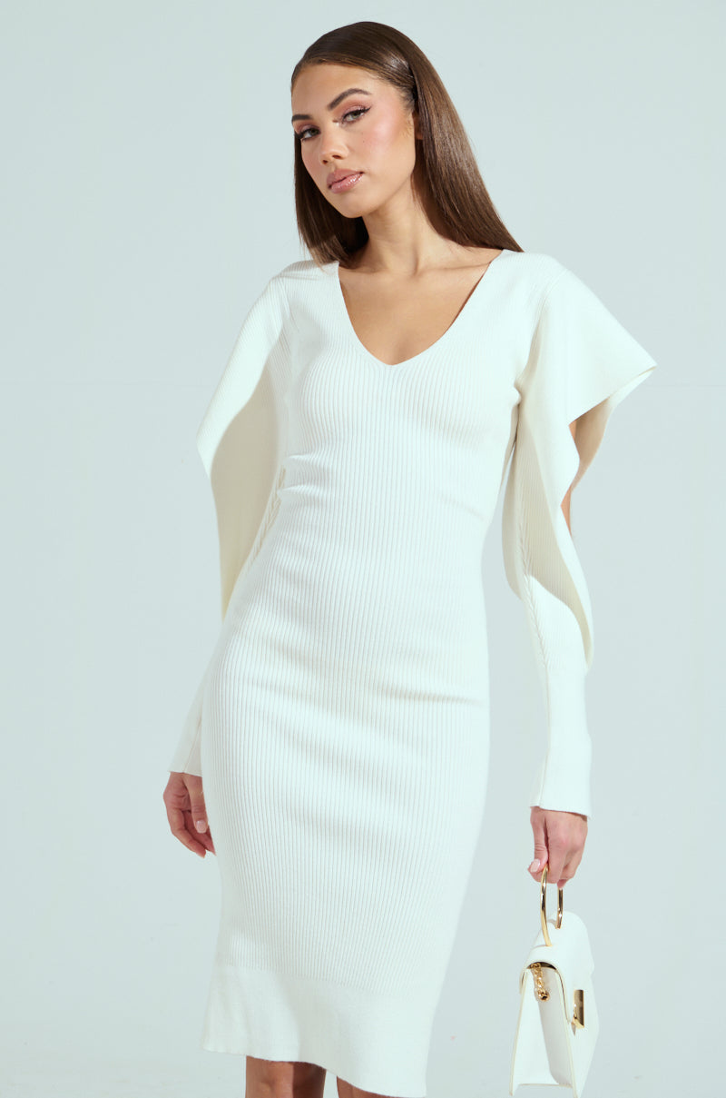 Dressed in the elegant OFF TO BRUNCH knit midi dress with a plunging V-neckline and unique open flap sleeves, she gracefully holds a small white handbag against a light gray backdrop.