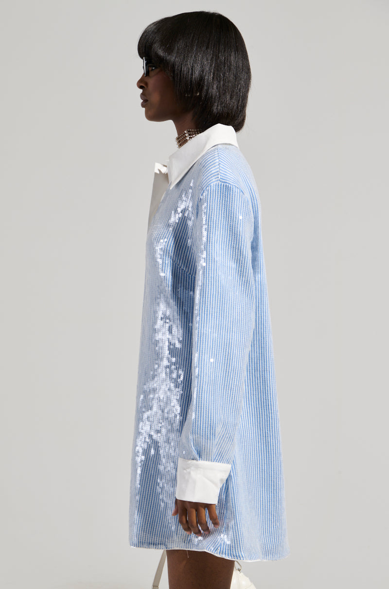 A woman with shoulder-length dark hair stands in profile, wearing the CHIC AF Sequin Poplin Mini Dress in light blue and white. The dress showcases a pinstripe pattern with an elegant white collar and cuffs, set against a plain and neutral background.