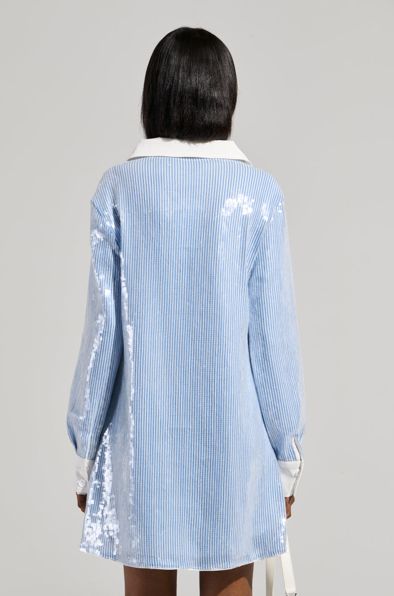 Rear view of a person with shoulder-length dark hair wearing the CHIC AF Sequin Poplin Mini Dress in a blue and white pinstripe pattern, featuring a crisp white collar and cuffs, standing against a plain background.