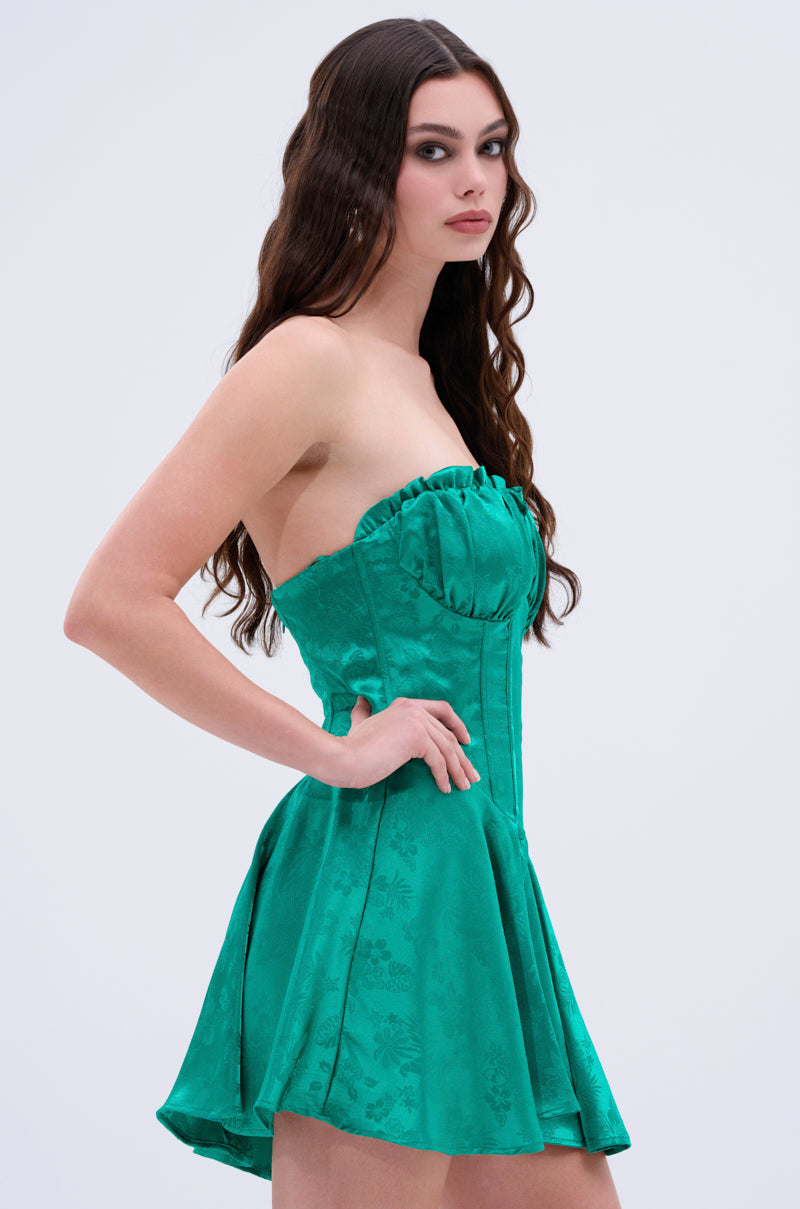 A woman with long wavy brown hair poses in the QUEEN OF IT ALL PRINTED SATIN MINI DRESS, a strapless emerald green mini dress with a skater silhouette and subtle floral print, set against a plain light background.