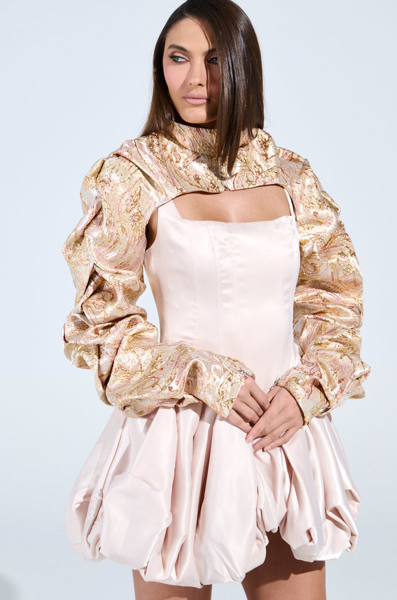 A woman in a soft pink dress with a structured bubble skirt and detailed, gold-patterned puff sleeves exudes party flair. The CORA BROCADE BOLERO would perfectly complement her look as she stands against a plain backdrop, glancing to the side with one hand resting on the other.