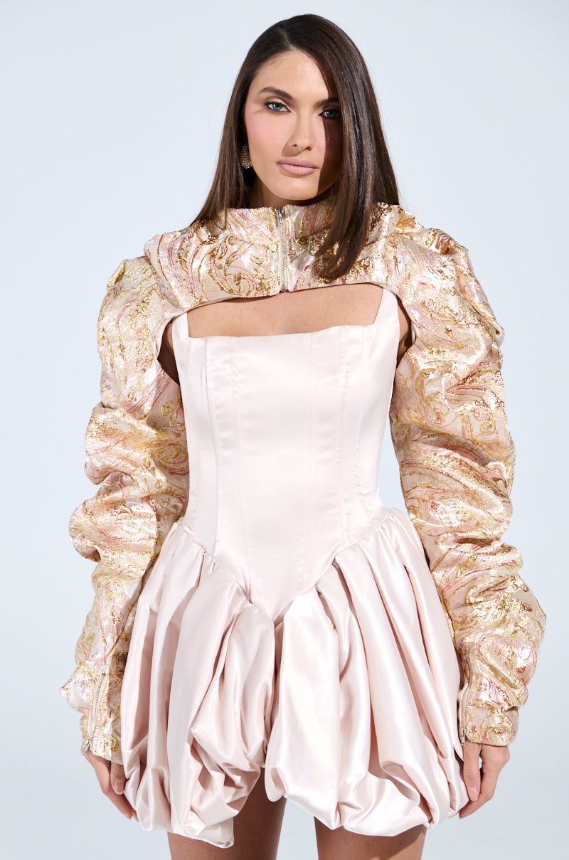 Someone with long brown hair is wearing the party-ready CORA BROCADE BOLERO, a stylish pale pink outfit with puffy, patterned sleeves over a voluminous skirt. The plain white background highlights this unique fashion piece.