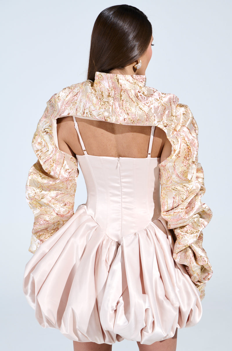 A woman with long brown hair elegantly wears the CORA BROCADE BOLERO, pairing it with a party-ready pale pink dress that features intricate textured design, voluminous puffy sleeves, a structured gathered skirt, back cutout, and fitted bodice with thin straps.