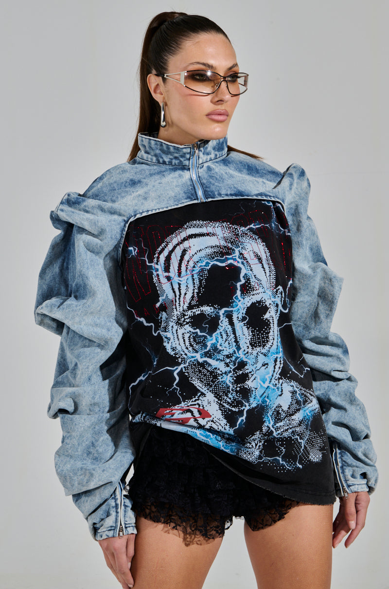 A woman with long hair tied back is wearing trendy sunglasses and the chic SPOIL ME DENIM BOLERO with exaggerated shoulders. Her top boasts an abstract graphic design, paired with a black textured skirt featuring ruched detailing. She poses confidently against a plain background.