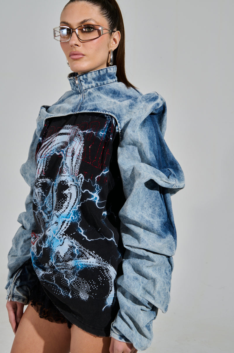 A woman with long brown hair and angular glasses models the bold SPOIL ME DENIM BOLERO, featuring a light wash, oversized sleeves, and ruched detailing. It's paired with a black graphic shirt with an intricate design as she stands against a plain background.