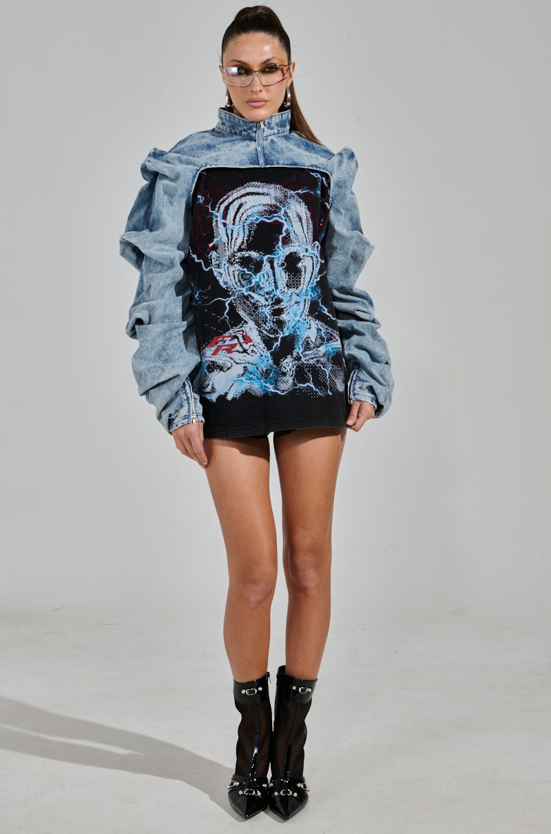 A model showcases the SPOIL ME DENIM BOLERO featuring a light wash with an abstract face design on the front, paired with a black mini skirt and black ankle boots. She completes her look with sunglasses while standing against a plain background.