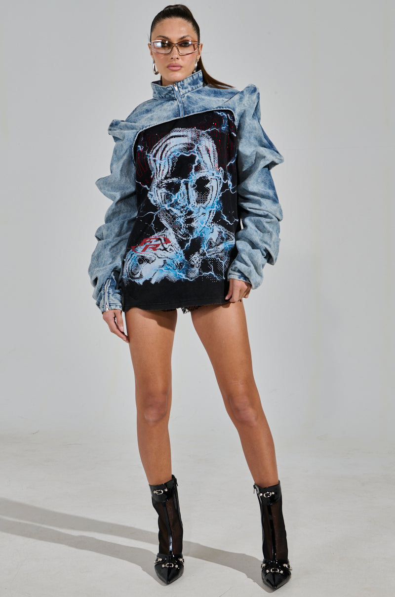 Sporting the SPOIL ME DENIM BOLERO with an abstract face design, a confident individual in sunglasses and black ankle boots stands in a minimalist setting. They have long hair tied back, black shorts, and the light wash denim adds casual elegance to their overall look.