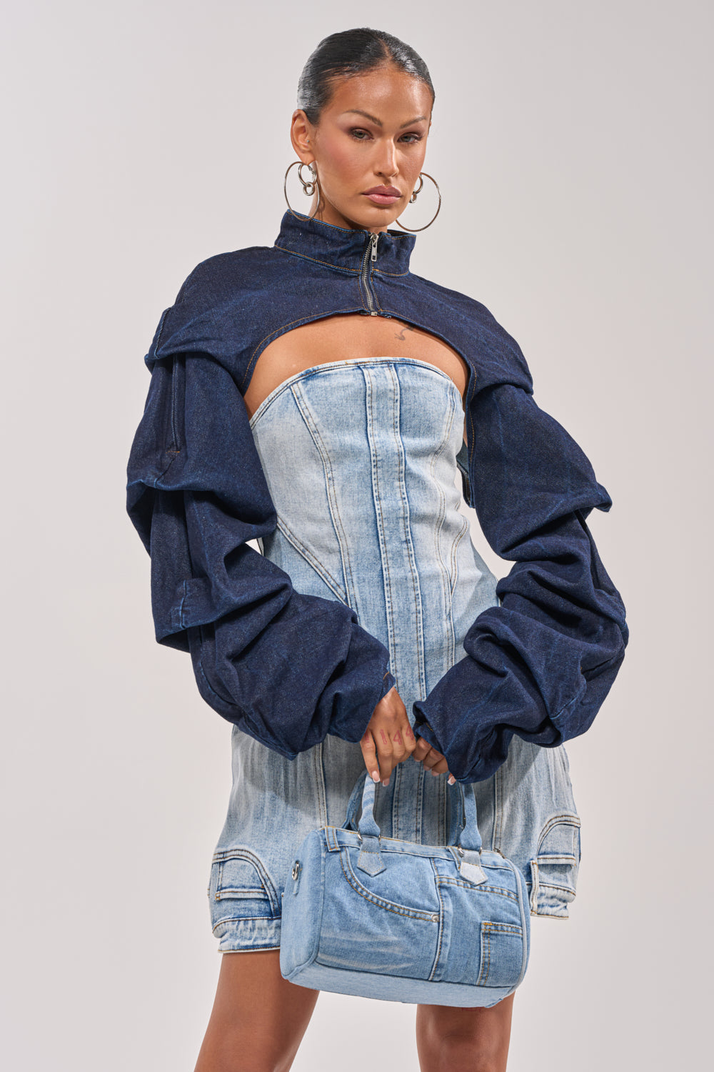 A woman exudes confidence in the SPOIL ME BOLERO IN DARK DENIM, styled over a strapless fitted denim dress. She completes her street-style look with a light blue denim handbag, large hoop earrings, and slicked-back hair. 0
