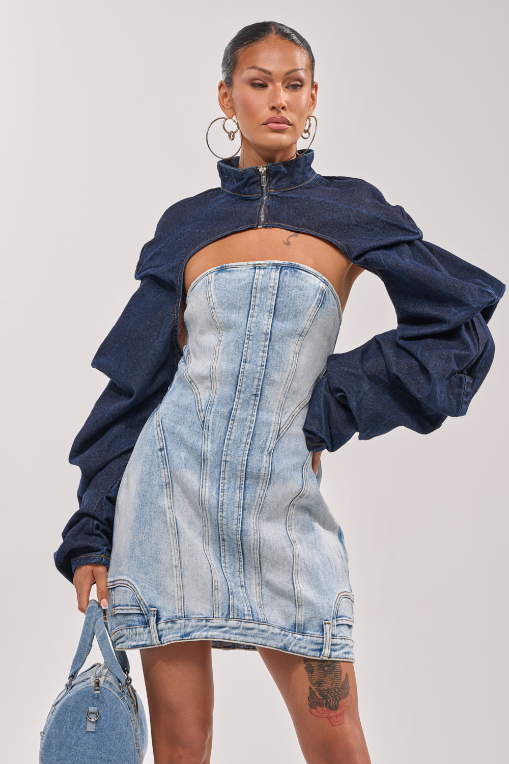 A woman poses confidently in the SPOIL ME BOLERO IN DARK DENIM, featuring dramatic dark blue sleeves, a zippered collar, and a light blue corset-style bodice. She accessorizes with a matching handbag and large hoop earrings. 2