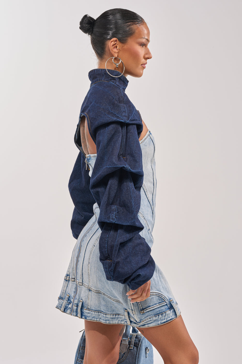 A woman with her hair in a bun and large hoop earrings wears the SPOIL ME BOLERO IN DARK DENIM—a cropped jacket with voluminous sleeves—layered over a light denim mini dress, standing in profile against a plain background. 3