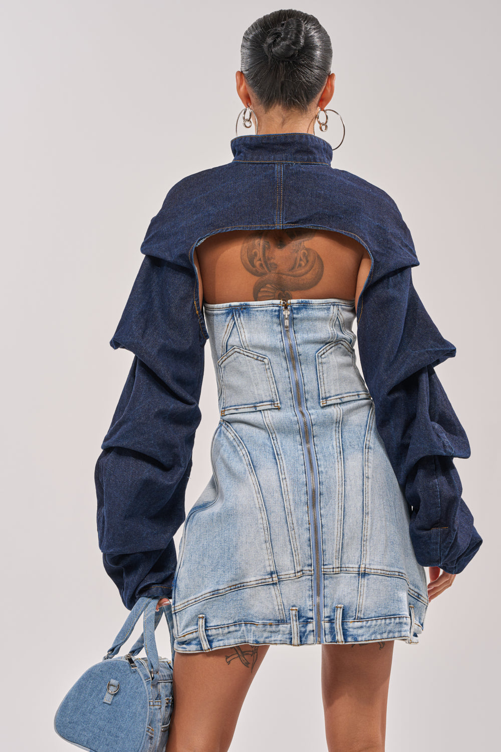 Seen from behind, a person with a bun and hoop earrings wears the SPOIL ME BOLERO IN DARK DENIM, featuring ruffled sleeves and an open back that reveals a tattoo. They're holding a blue denim handbag. 4