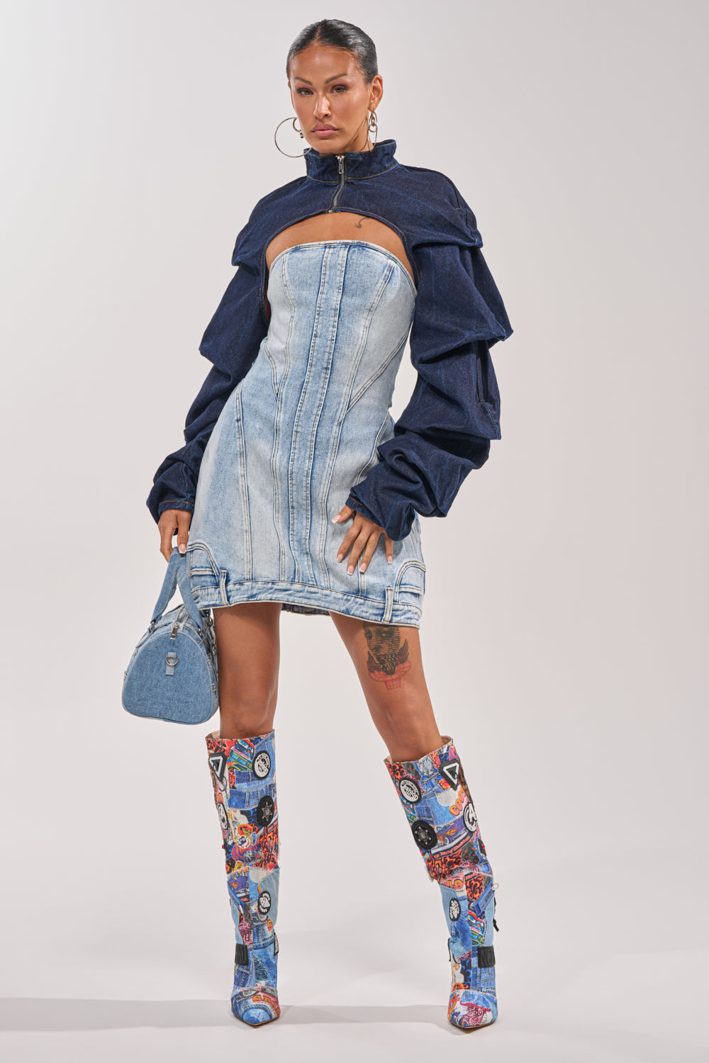A woman poses in the Spoil Me Bolero in Dark Denim, paired with a strapless mini dress, denim handbag, and tall patchwork boots. With hoop earrings and her hair pulled back, she epitomizes modern street-style cool. 5