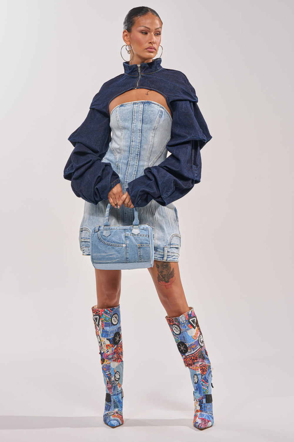 A woman poses against a plain background in a strapless light denim mini dress, the SPOIL ME BOLERO IN DARK DENIM with puffy sleeves, a matching denim handbag, and knee-high boots detailed with colorful patchwork street style. 6