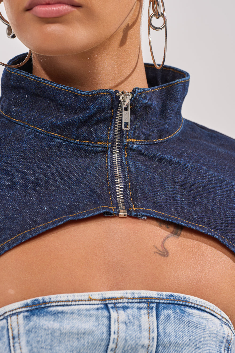 Close-up of a woman wearing the SPOIL ME BOLERO IN DARK DENIM with a zipper collar over a light blue bandeau top, accessorized with large hoop earrings and showing a small chest tattoo for a bold street-style look. 7