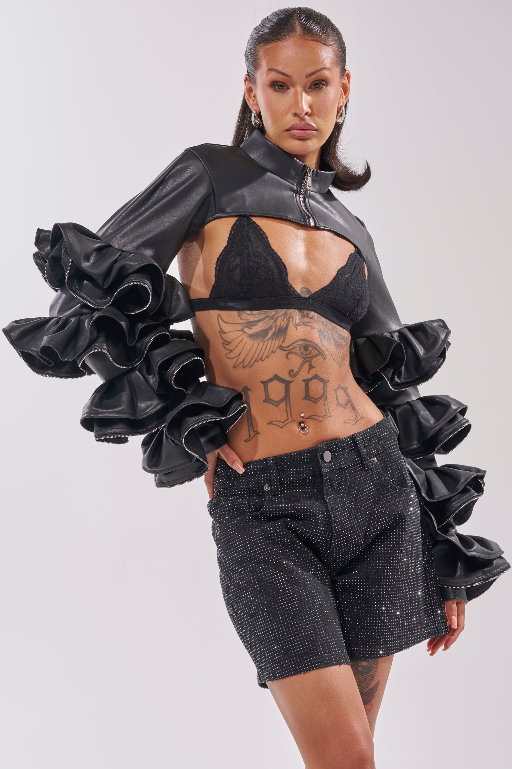 A woman with tattoos and styled dark hair poses confidently in the DON'T MESS RUFFLE BOLERO—a cropped black jacket with dramatic ruffled sleeves—paired with a black lace bra and sparkly black shorts. 0