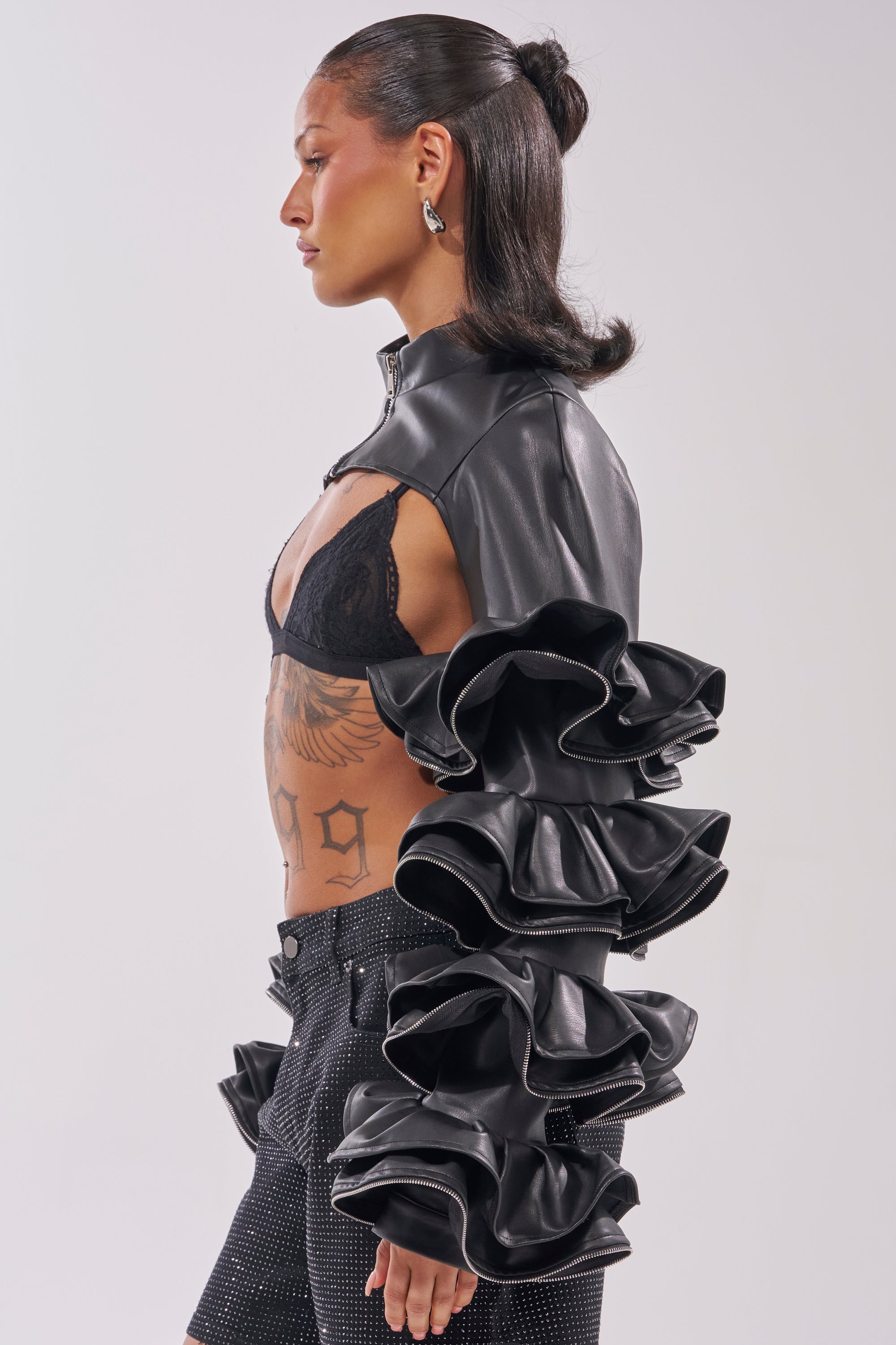 A person in profile wears the DON'T MESS RUFFLE BOLERO—a cropped faux leather jacket with dramatic ruffled sleeves—paired with a black lace bra and sparkly black shorts. Visible tattoos adorn their torso, and their dark hair is in a chic half-updo. 2