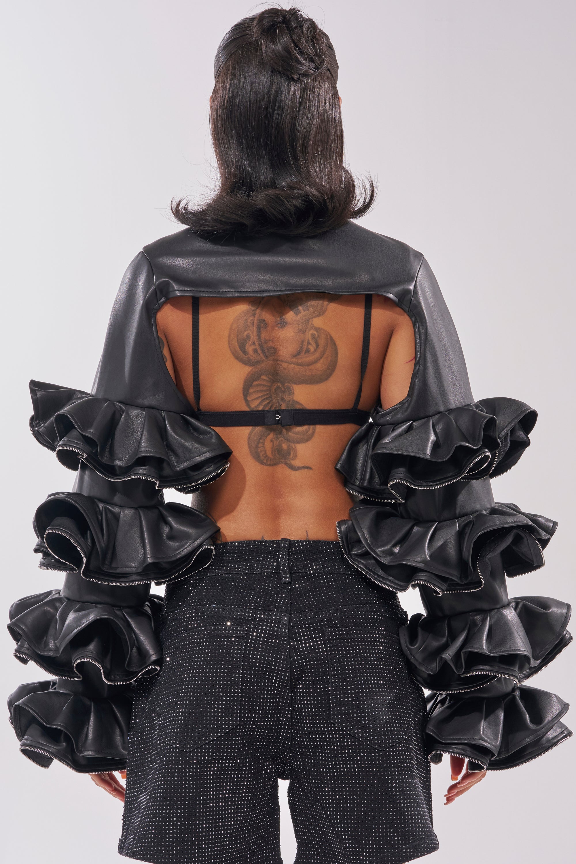 A person is seen from behind in soft waves, wearing the DON'T MESS RUFFLE BOLERO with dramatic ruffled sleeves and sparkly black shorts, showing off a large upper-back tattoo. 3