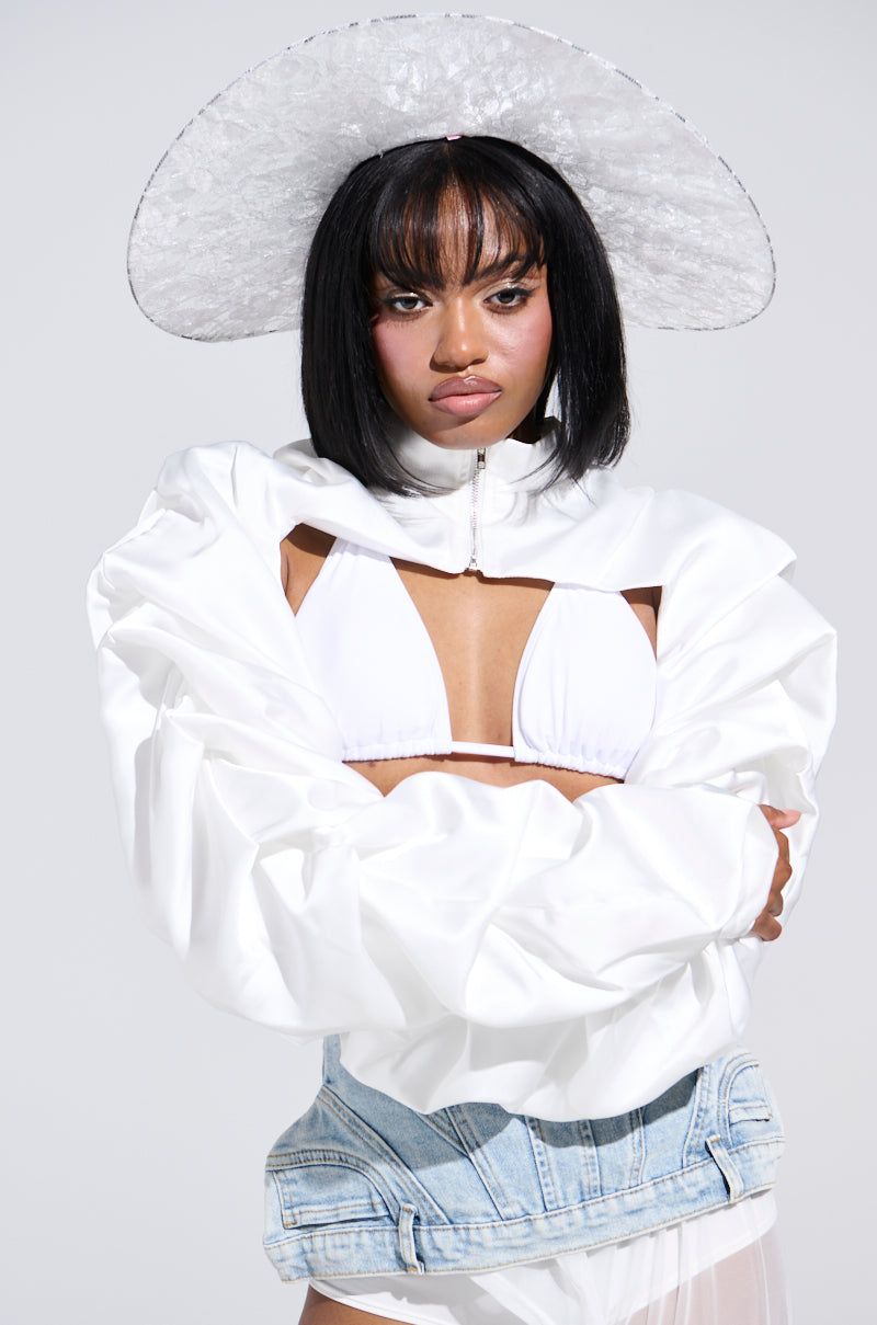 A woman confidently poses in the chic Cora Satin Bolero in white with a wide-brimmed hat and bikini top. Her short black hair with bangs complements light blue denim jeans as she crosses her arms against a plain background.