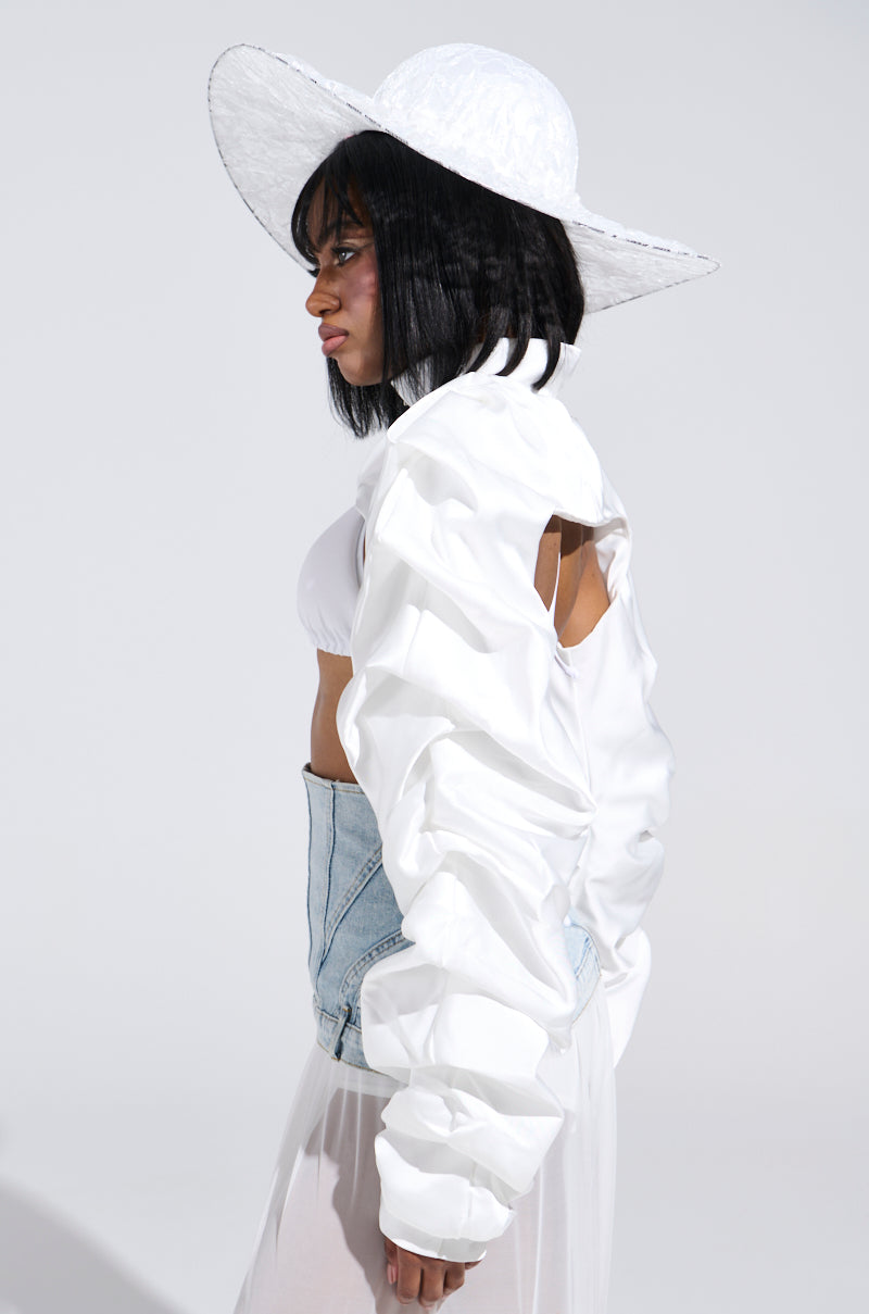 A person stands in profile against a plain backdrop, wearing the chic CORA Satin Bolero in white. This ensemble features a large white wide-brimmed hat, voluminous jacket with a metallic front zip closure, denim corset, and is elegantly finished with a light, semi-transparent skirt.