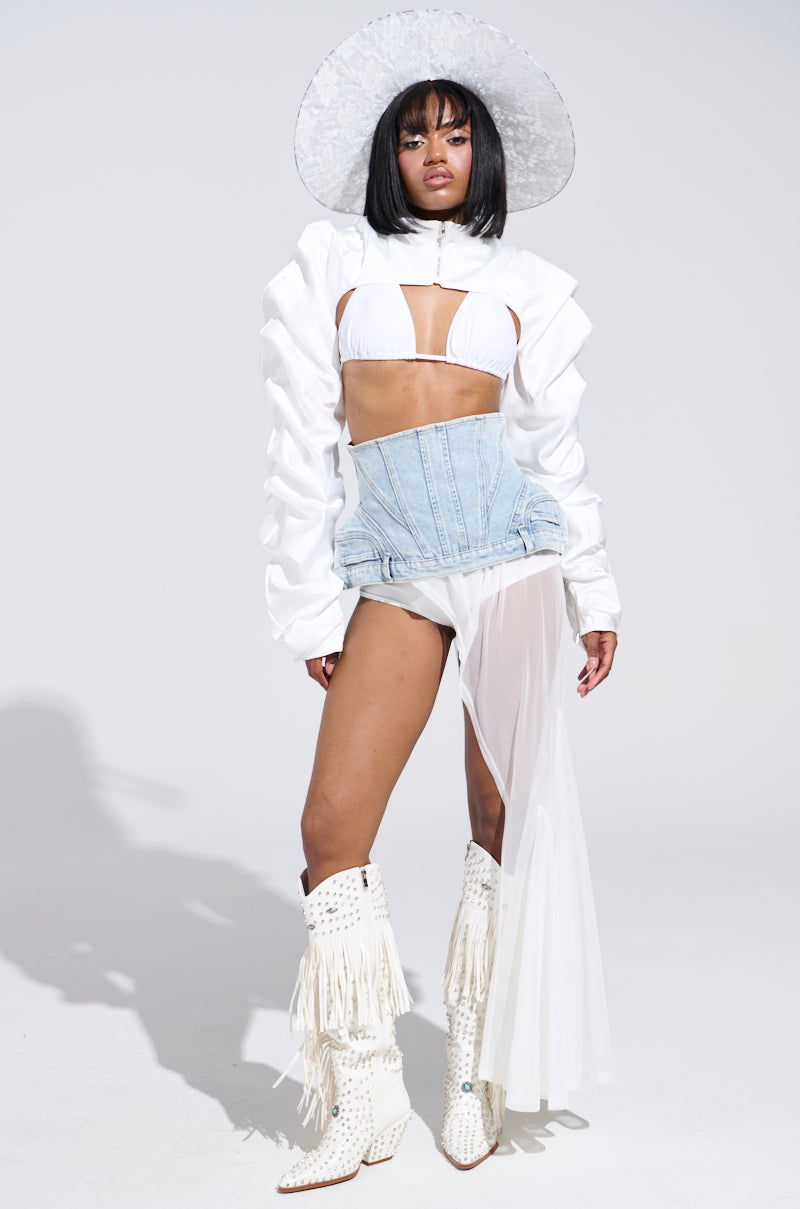 Dressed in the CORA SATIN BOLERO IN WHITE, the person poses stylishly in a chic ensemble with a wide-brimmed hat, white bikini top, denim corset, and sheer skirt. Knee-high boots with fringe detailing complement the look against a plain white backdrop. A metallic front zip closure completes it.