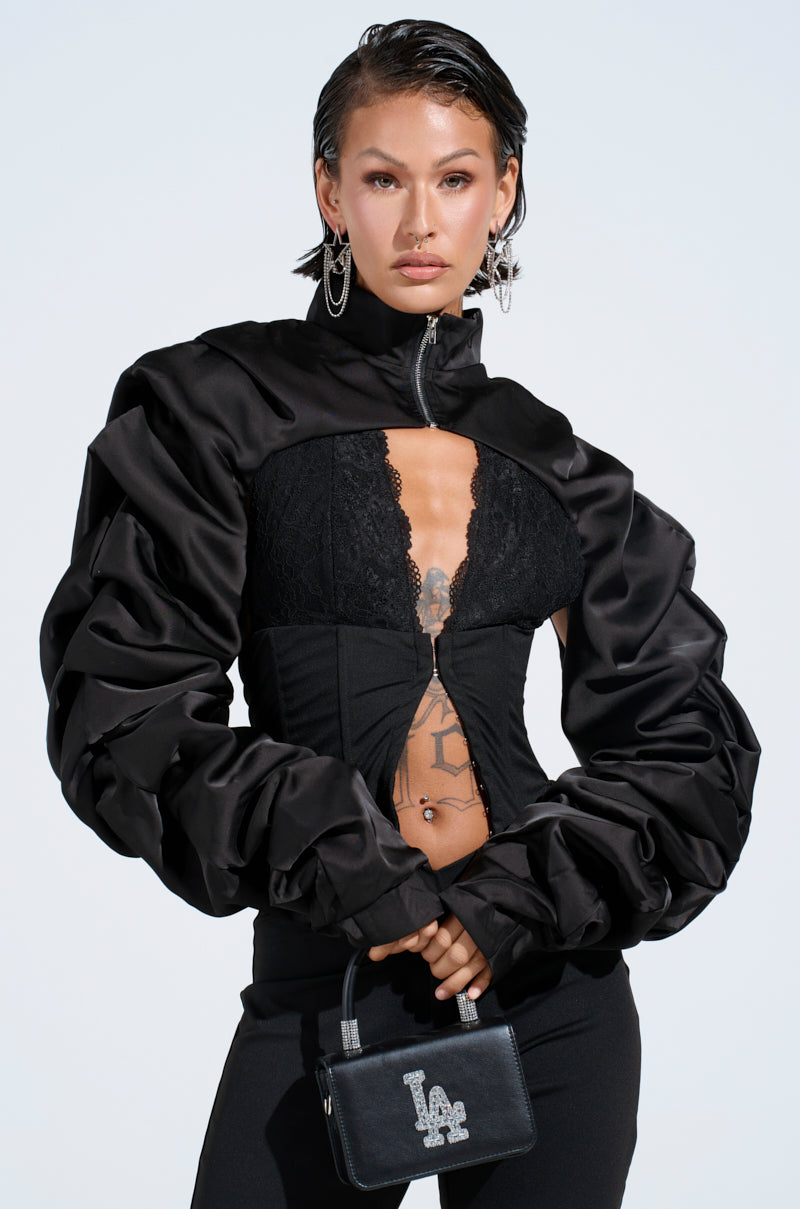 A person poses against a plain background wearing the CORA SATIN BOLERO IN BLACK, featuring voluminous sleeves, over a lace top and black flared leather pants. They hold a small handbag with "LA" in rhinestones and have short hair with large earrings.