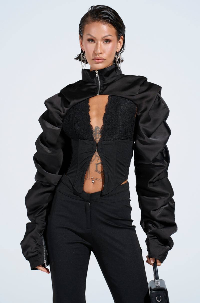 A person with wet-look hair wears a stylish black outfit featuring the CORA SATIN BOLERO IN BLACK jacket, alongside a lace top and flared leather pants. They accessorize with a small handbag, hoop earrings, and a necklace against a plain white background.