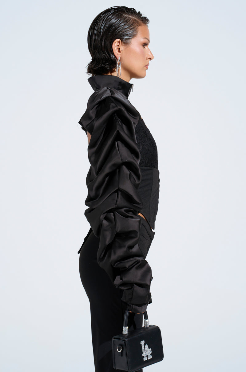 A person in profile wears a black, satin bolero with ruffled long sleeves and flared leather pants against a plain background. They carry a small black handbag featuring "L.A." and have slicked-back hair complemented by dangling earrings.