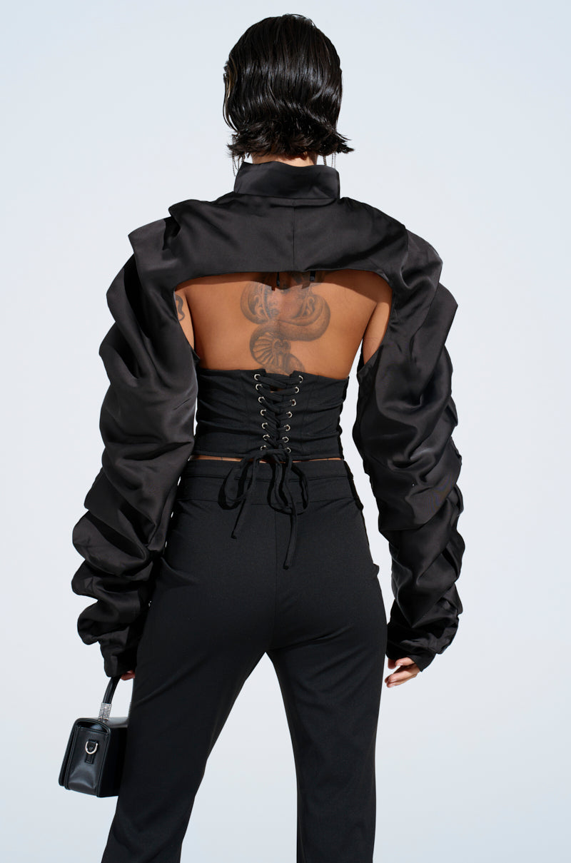 A person showcases the Cora Satin Bolero in black with open back and lace-up detail, paired with flared leather pants. They carry a small black handbag, and tattoos stand out against the light grey background, enhancing their edgy style.