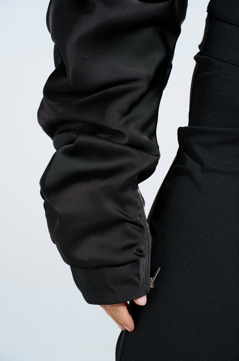A close-up of someone wearing the Cora Satin Bolero in black reveals its ruched sleeve, adorned with a visible zipper near the wrist, exuding flamboyant elegance against a plain white backdrop.