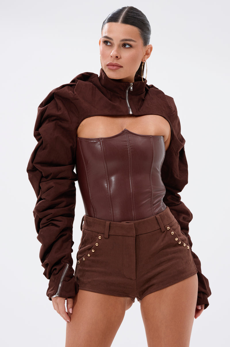 A woman poses confidently against a plain background wearing the RILEY STACKED FAUX SUEDE BOLERO, a dark brown structured jacket with ruched sleeves, paired with matching high-waisted shorts featuring gold buttons. 0