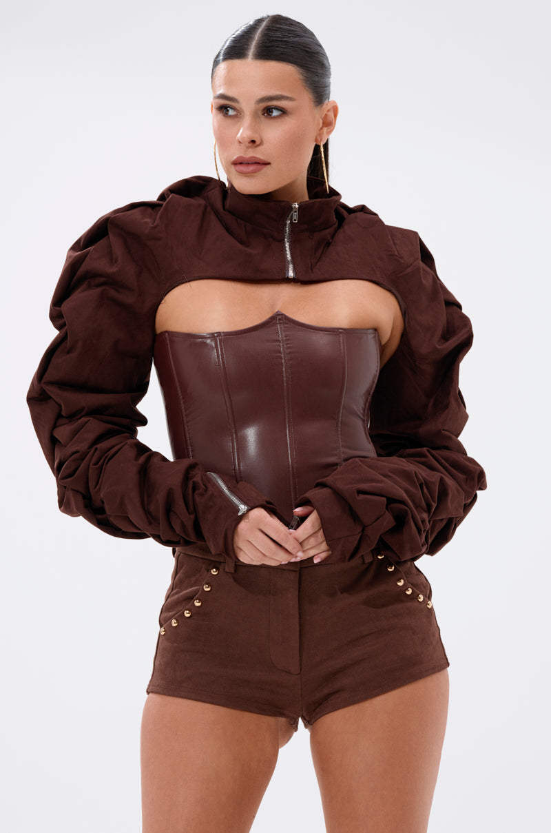 A woman stands against a plain background wearing the RILEY STACKED FAUX SUEDE BOLERO, a dark brown off-shoulder cropped jacket with puffy sleeves, styled with a structured brown corset and high-waisted shorts featuring gold buttons. 1