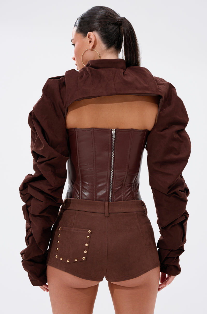 A woman with a ponytail is seen from the back wearing the RILEY STACKED FAUX SUEDE BOLERO, a brown, long-sleeve open-back jacket, paired with a corset-style leather top and high-waisted brown shorts with gold stud details. 4