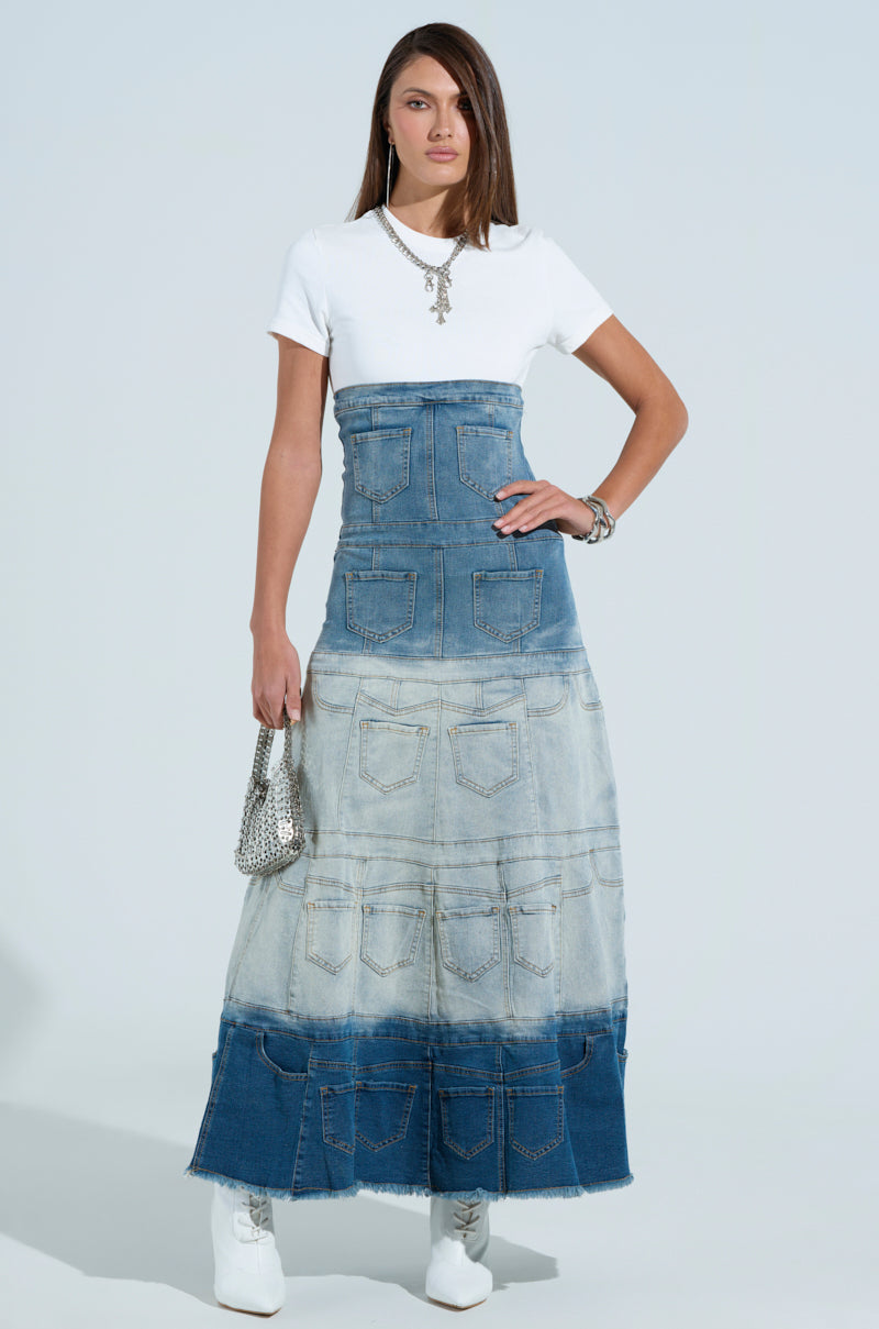 Against a light background, a person wears the DENIM ON DENIM LAYERED MAXI DRESS with multiple pockets, paired with a white T-shirt and boots. They accessorize with a small silver handbag, necklace, and bracelet.
