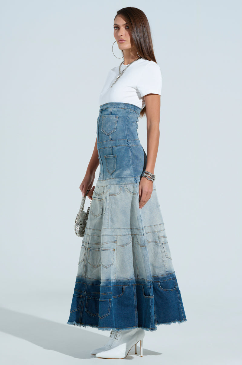 A woman stands against a light background wearing the Denim On Denim Layered Maxi Dress, accessorized with a silver purse, hoop earrings, and bracelets. Her long straight hair and white heeled boots complete the look perfectly.