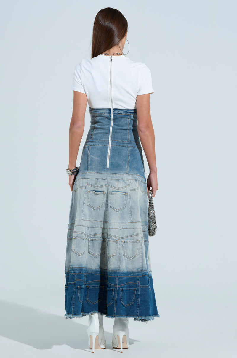 A person with long hair, facing away, wears a white top paired with an ombre denim skirt boasting blue shades and diverse pocket designs. They hold a small bag and wear white boots, completing the chic DENIM ON DENIM LAYERED MAXI DRESS ensemble.
