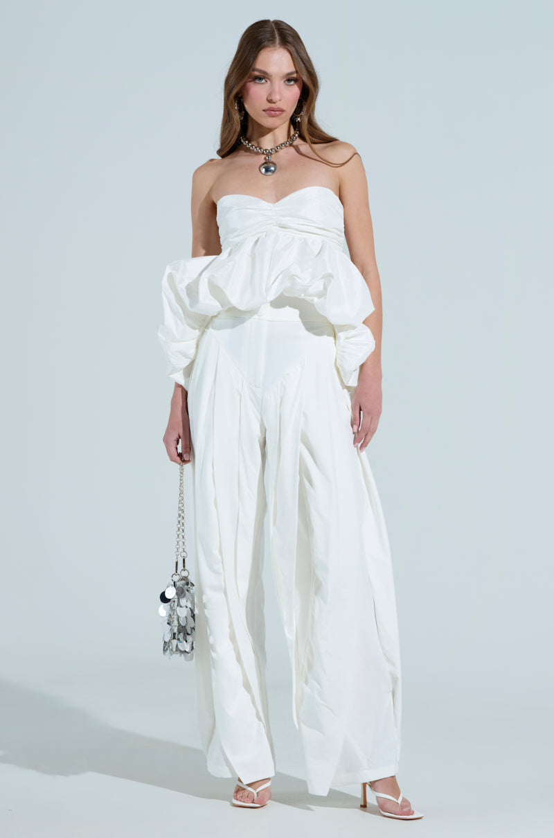A woman poses against a plain backdrop, wearing the AMBER WIDE LEG PLEATED TROUSER PANT IN WHITE with a strapless voluminous white top. Her look is completed with a chunky necklace, a handbag with metallic details, and open-toed heels.