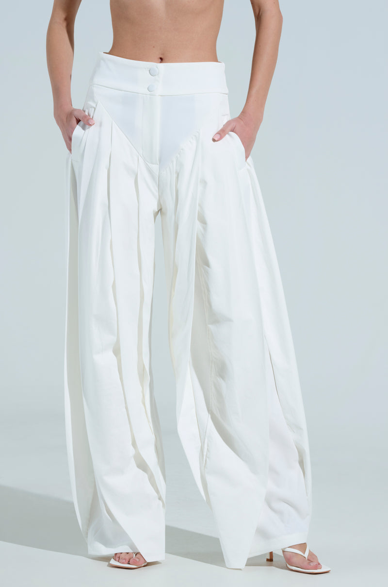 A person in AMBER WIDE LEG PLEATED TROUSER PANT IN WHITE stands casually, hands tucked in pockets. They're barefoot except for white flip-flops, against a plain background.