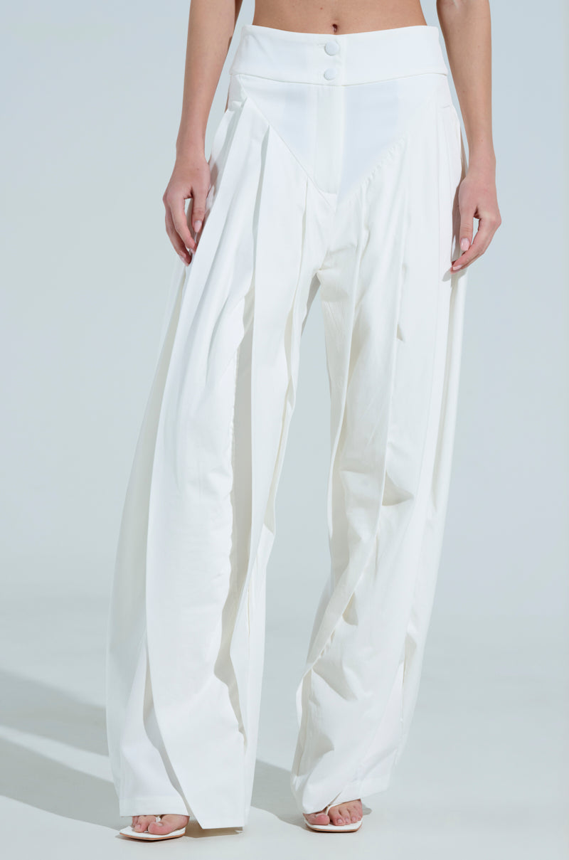 A person wearing the AMBER WIDE LEG PLEATED TROUSER PANT IN WHITE stands against a light backdrop, showcasing its high-rise pleated design with a button closure. Their feet appear in open-toed shoes.