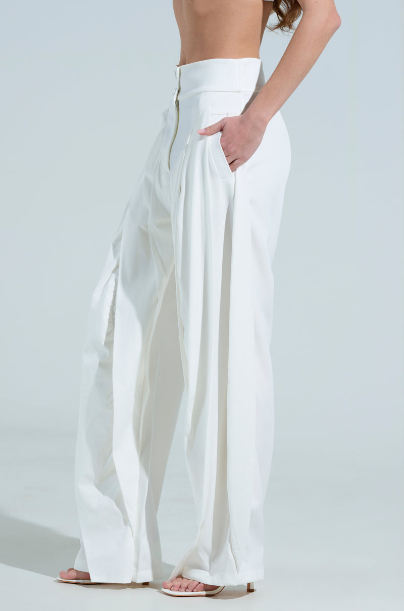 A person is wearing the AMBER WIDE LEG PLEATED TROUSER PANT IN WHITE, featuring a high-rise waistband and pockets, paired with heeled sandals. The side view showcases the flowy fabric and relaxed fit against a light background.