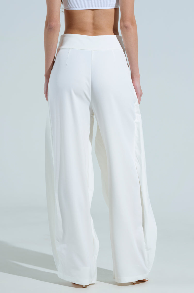 A person is standing with relaxed hands, wearing the AMBER WIDE LEG PLEATED TROUSER PANT IN WHITE and a white crop top. The background is a plain, light color.