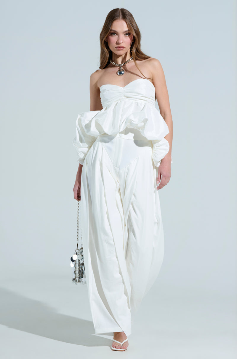 A woman in the AMBER wide-leg pleated trousers and a flowing strapless ruffled top accessorizes with a silver necklace and carries a metallic handbag, standing confidently against a light gray background.