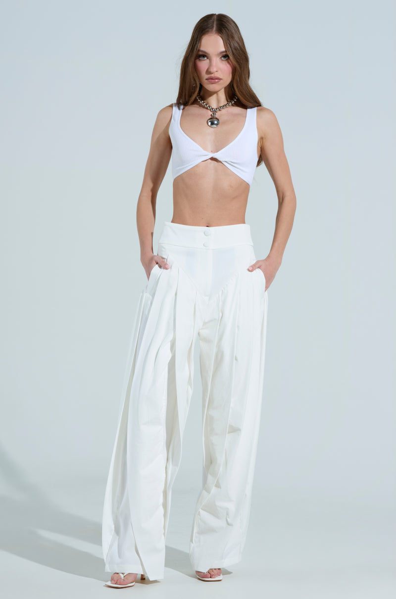 A person with long hair wears a white cropped top and AMBER wide-leg pleated trousers in white, featuring a high-rise waistband. The look is elegant with a round pendant necklace, standing against a plain light background, hands resting casually in pockets.