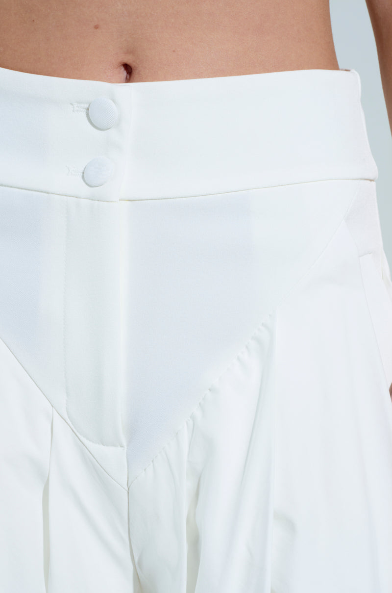 A person wears the AMBER WIDE LEG PLEATED TROUSER PANT IN WHITE, which features a high rise waistband with buttons and pleats for an elegant texture. The background is soft and neutral.